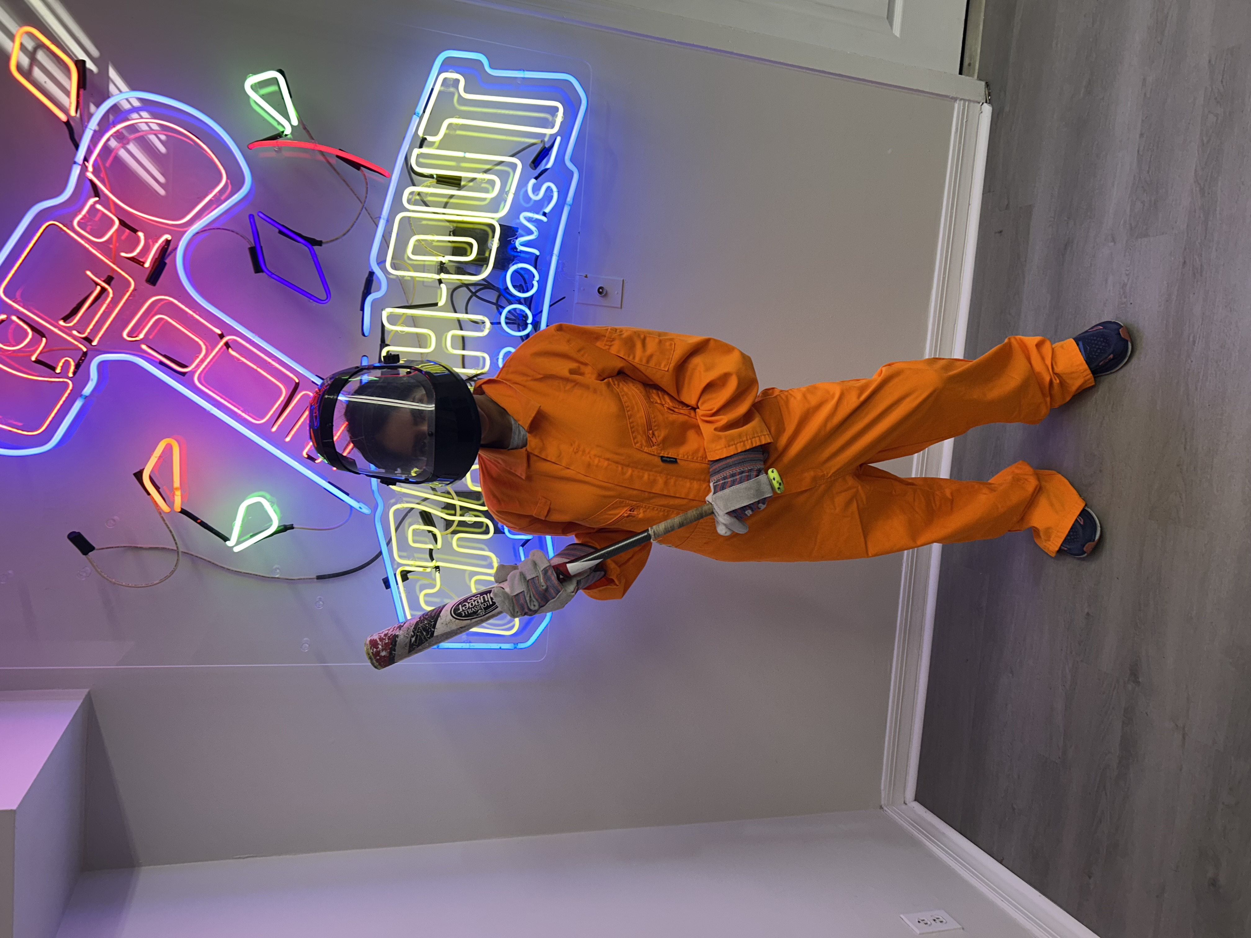 Crash Out customer in orange jumpsuit posing in front of neon Crash Out Rage Rooms sign