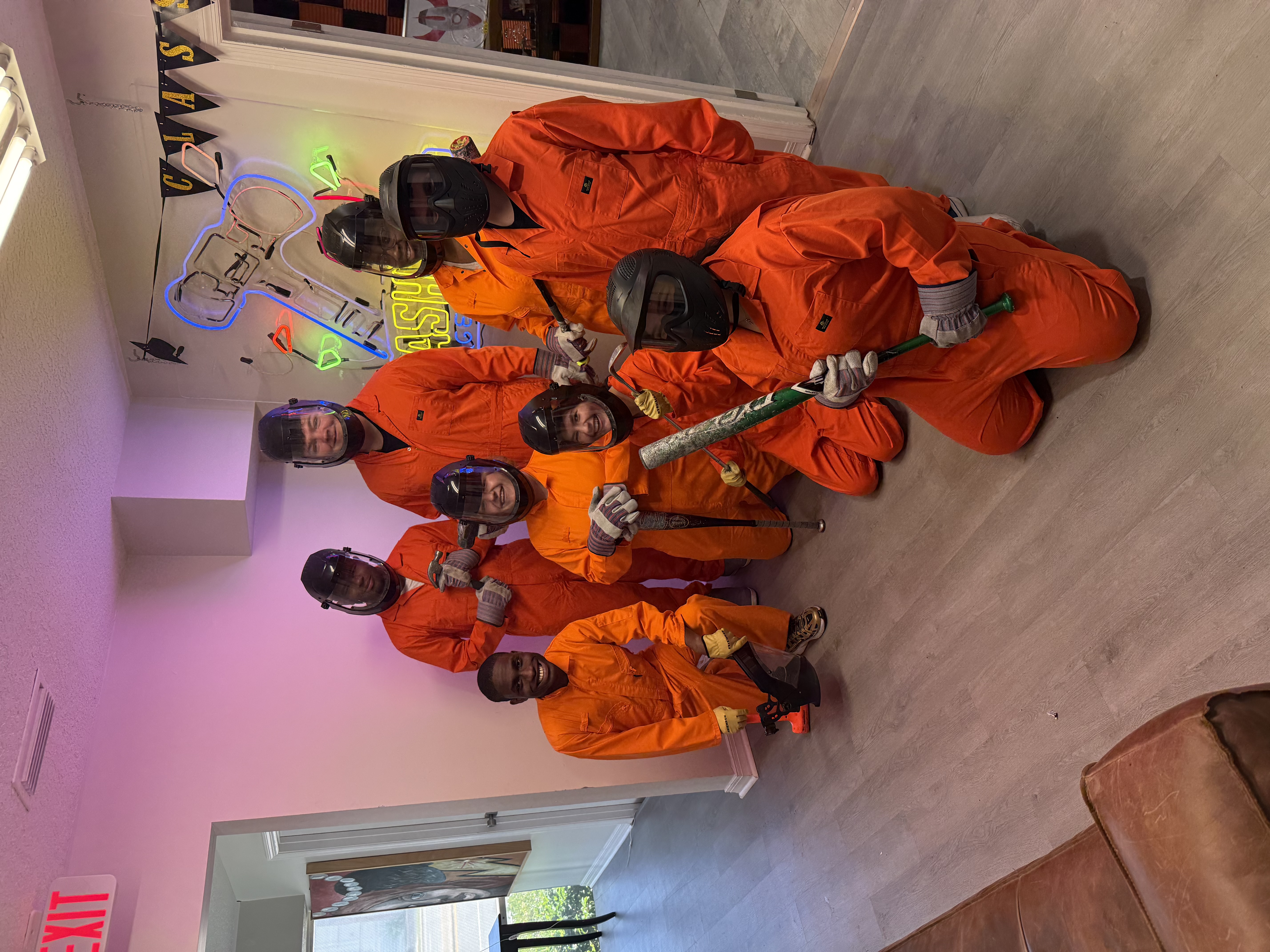 Group of eight Crash Out Rage Rooms customers in orange jumpsuits and protective gear holding bats and tools