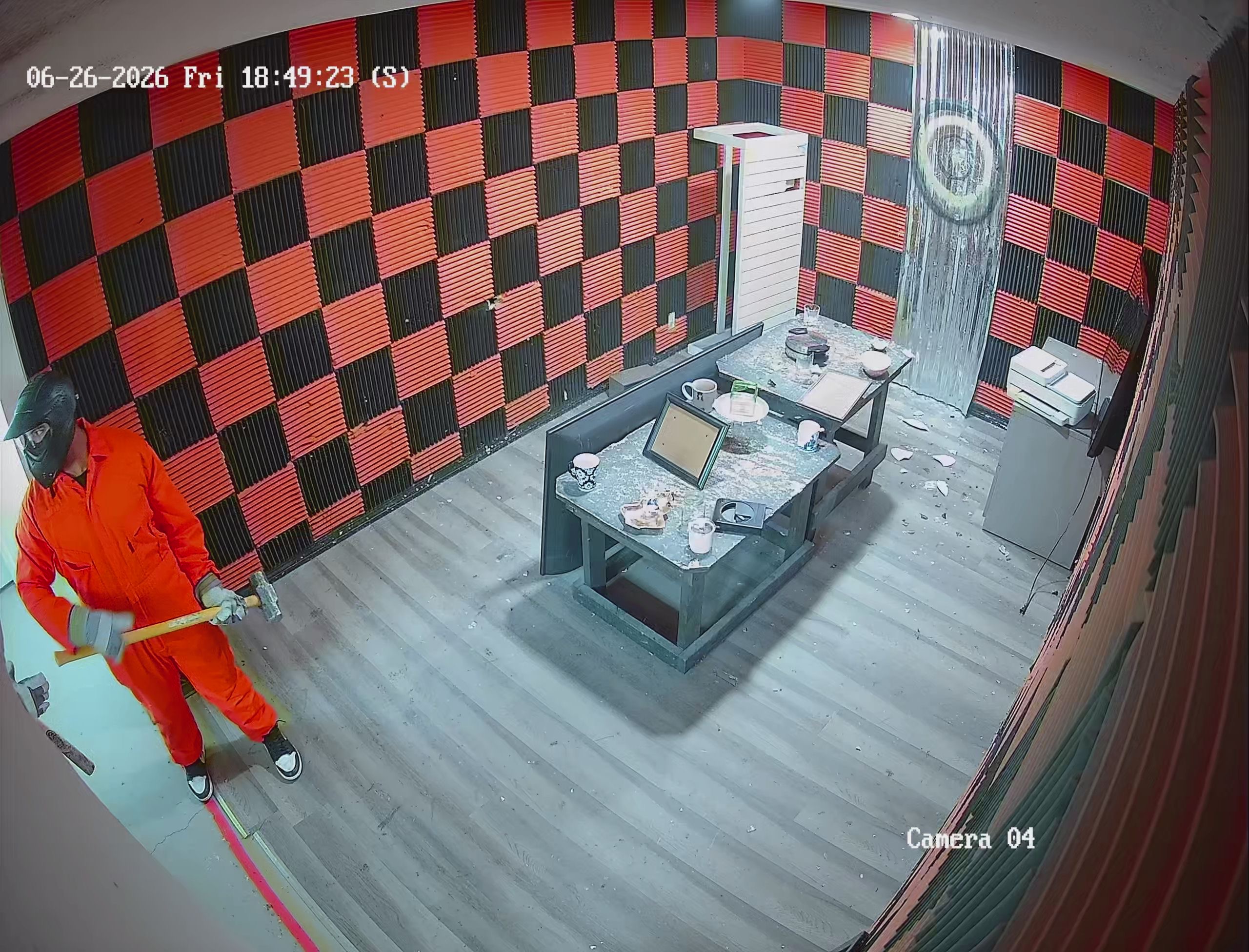 Security camera view of guest in orange jumpsuit mid-smash inside Crash Out rage room with red and black checkered walls